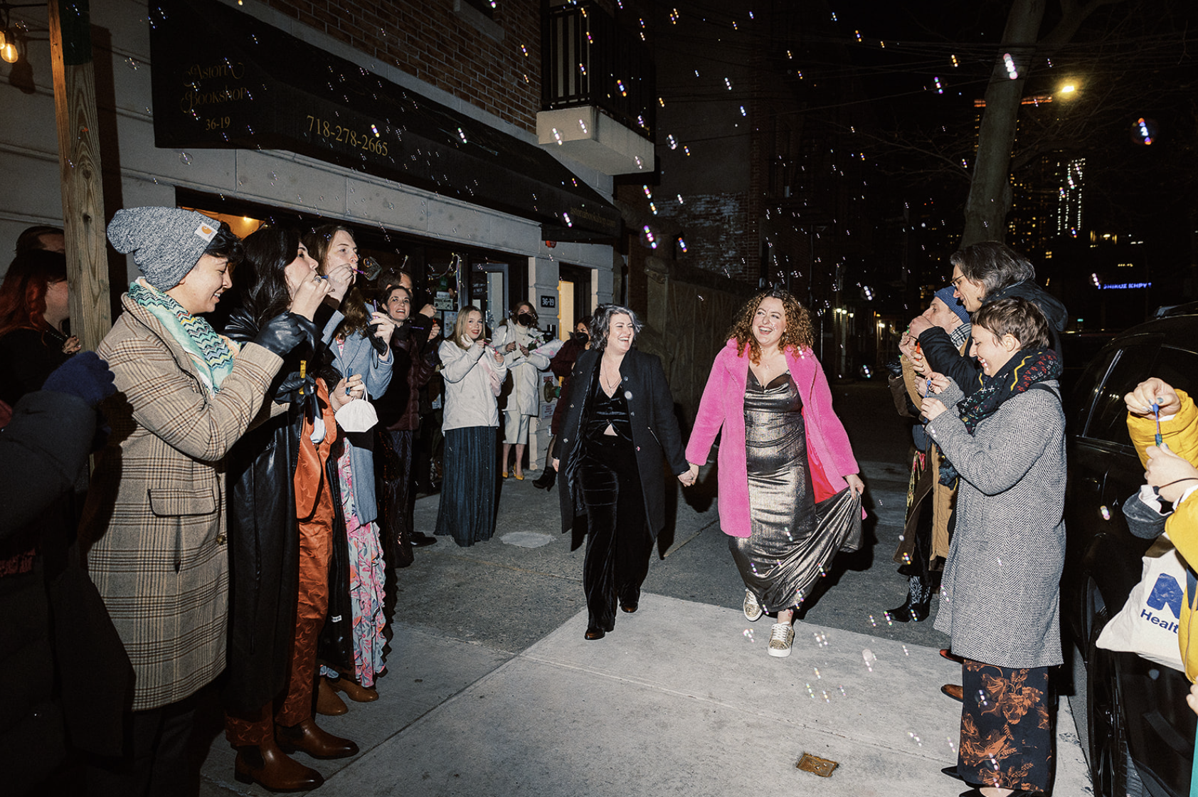 meg and jeanna walk on a ny street holding hands, surrounded by guests in winter coats blowing bubbles and smiling