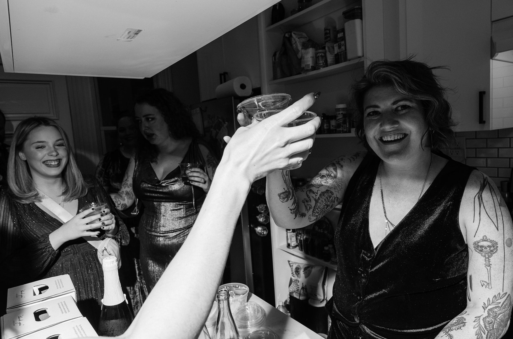 b&w photo of meg standing in kitchen smiling, clinking a glass of wine with another person, while jeanna and other guests talk nearby