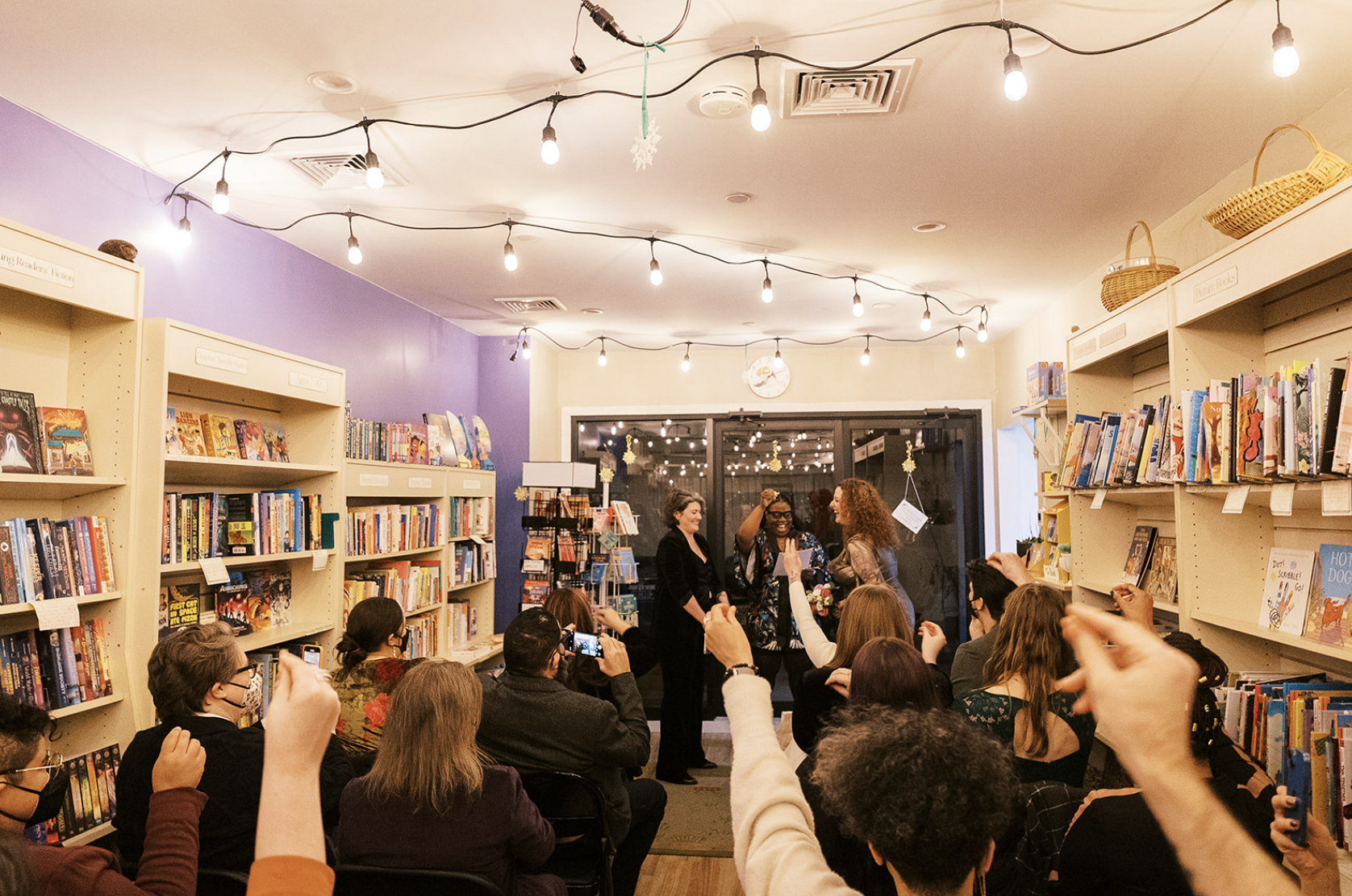 interior of bookshop with warm wooden bookshelves lining either side of the room, meg and jeanna standing in front with their officiant mecca woods, and seated guests facing them with fingers up snapping