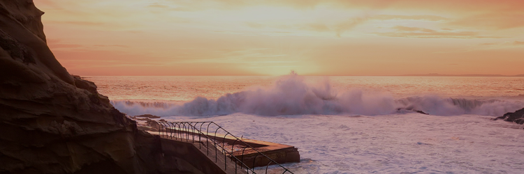 ocean waves crashing on beach with cliff and outdoor stairs in the foreground, sunset behind